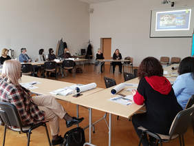 W sali która ma jasne ściany, jasno brązową podłogę, przy stołach ułożonych w kształcie litery u odbywa się spotkanie. Uczestnicy, widzimy 10 osób siedzą przy stołach. Przy osobny stole siedzą dwie panie prelegentki.