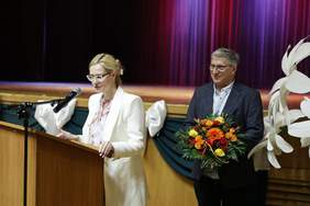 Pokaż zdjęcie: Kobieta w jasnym garniturze przemawia przy mównicy z mikrofonem na tle sceny. Obok niej stoi mężczyzna w ciemnej marynarce, trzymający bukiet z pomarańczowych i żółtych kwiatów.