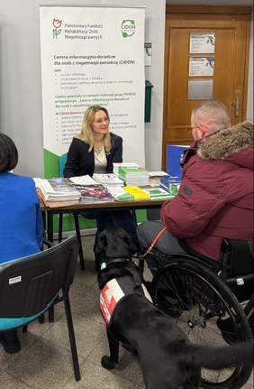 Pokaż zdjęcie: Na zdjęciu siedzi kobieta z jasnymi włosami, uśmiechnięta, ubrana w jasny bluzkę i ciemną marynarkę. Na stole przed nią leżą ulotki, broszury i książeczki ułożone w stosy. Za stołem stoi pionowy roll-up z logo i tekstem informacyjnym (Centrum informacyjno-doradczego dla osób z niepełnosprawnością, zwanym również CIDON). Widoczne są też logo Państwowego Funduszu Rehabilitacji Osób Niepełnosprawnych. Kobieta udziela informacji panu, który siedzi na wózku i do niego jest przypięty pies przewodnik. Osoba na wózku ma czerwoną pikowaną kurtkę z futrzanym kapturem i krótkie jasne włosy. 