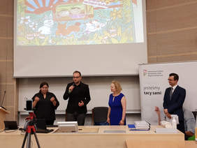 Pokaż zdjęcie: od lewej kobieta na caarno ubrana tłumaczy na język migowy, obok mężczyzna w garniturze mówi do mikrofonu, obok kobieta, blondynka w niebieskiej sukience, całkiem z prawej na tle baneru PFRON mężczyzna w garniturze 