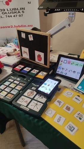 Pokaż zdjęcie: Na stole leżą różne urządzenia takie jak laptopy, tablety, urządzenia te wspierają mowę oraz piktogramy ułatwiające komunikację
