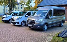 Pokaż zdjęcie: Trzy zaparkowane vany stojące obok siebie na wybrukowanym placu. Dwa z nich to białe samochody Renault, a trzeci to szary Ford z podwyższonym dachem. W tle widać budynek z żółtymi ścianami i drzewa. 