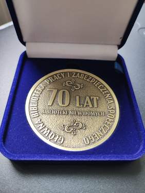 Pokaż zdjęcie: W niebieskim pudełku znajduje się złoty medal. Medal z okazji 70-lecia Biblioteki Niewidomych, Główna Biblioteka Pracy i Zabezpieczenia Społecznego