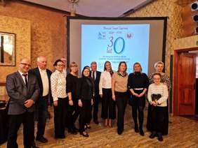 Pokaż zdjęcie: Grupa trzynastu osób stoi w sali restauracyjnej na tle dużego ekranu. Wyświetlany jest na nim slajd informujący o jubileuszu 30-lecia WTZ w Kaliszu prowadzonego przez Fundację Inwalidów i Osób Niepełnosprawnych „Miłosierdzie”.