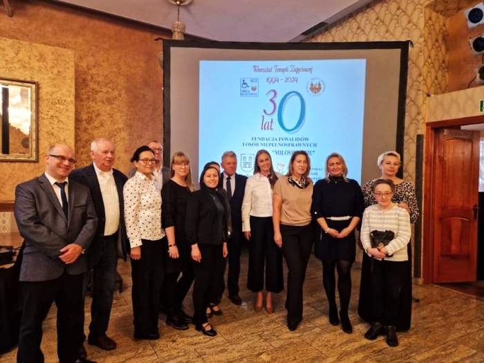Pokaż zdjęcie: Grupa trzynastu osób stoi w sali restauracyjnej na tle dużego ekranu. Wyświetlany jest na nim slajd informujący o jubileuszu 30-lecia WTZ w Kaliszu prowadzonego przez Fundację Inwalidów i Osób Niepełnosprawnych „Miłosierdzie”.