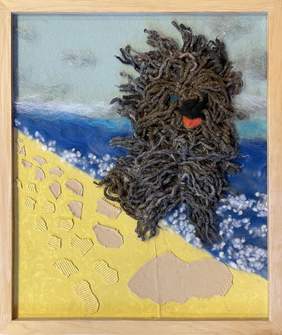 Pokaż zdjęcie: Na żółtej plaży siedzi pies wykonany z brązowej i szarej wełny. W tle niebieska woda wykonana z wełny. W górnej części tła niebo w kolorze szarym z szarymi chmurami.