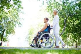 Pokaż zdjęcie: W parku znajduje się kobieta i mężczyzna. Pan siedzi na wózku inwalidzkim, a pani go prowadzi. W tle widać zielone drzewa i światło słoneczne. 