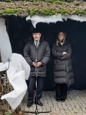 Pokaż zdjęcie: Pod szopką stoi mężczyzna, który przemawia do mikrofonu. Składa zebranym świąteczne życzenia. Obok niego stoi kobieta. Po lewej stronie widać dekoracje imitujące śnieg. 