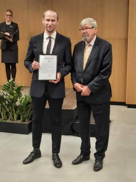 Pokaż zdjęcie: W sali stoi dwóch mężczyzn, którzy pozują fotoreporterom. Jeden z panów prezentuje dyplom. W tle widać kobietę. 