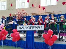 Pokaż zdjęcie: Na scenie trwa występ artystyczny w wykonaniu zespołu Świniczanki. Kobiety i mężczyźni ubrani są na ludowo. Panie śpiewają, a panowie grają na instrumentach. 