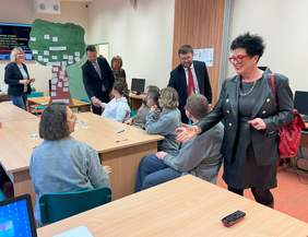 Pokaż zdjęcie: Grupa osób siedząca przy stole w trakcie zajęć edukacyjnych, jednej z osób Prezes PFRON podaje rękę na przywitanie, w tle widzimy tablice na ścianie