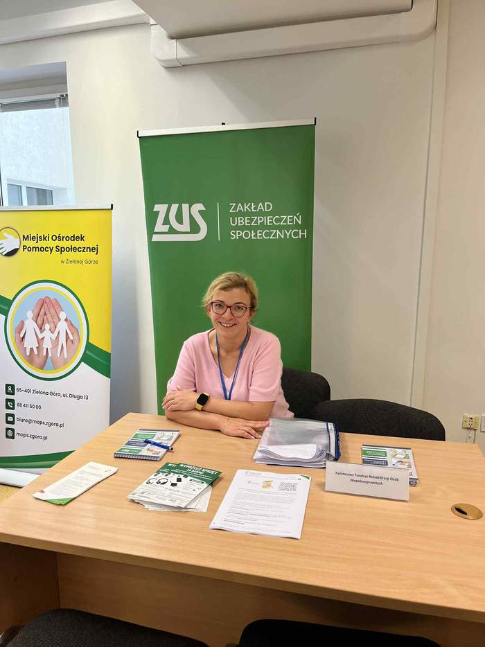 Pokaż zdjęcie: Uśmiechnięta kobieta ubrana w różowy sweter, znajdująca się przy biurku z materiałami PFRON oraz na tle baneru z logo ZUS