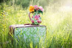 Walizka ukryta w trawie,"skąpanej"w słońcu, a na niej stoi kolorowy wazon z kwiatami