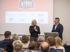 Pokaż zdjęcie: Zdjęcie przedstawia kobietę przemawiającą do mikrofonu podczas oficjalnego wydarzenia. Obok niej stoi mężczyzna przy mównicy. W tle widoczne jest logo klubu sportowego "Start Białystok" oraz logotypy partnerów, takich jak PFRON, Ministerstwo Sportu i Turystyki, TVP3 Białystok i inne. Na pierwszym planie siedzą uczestnicy wydarzenia.