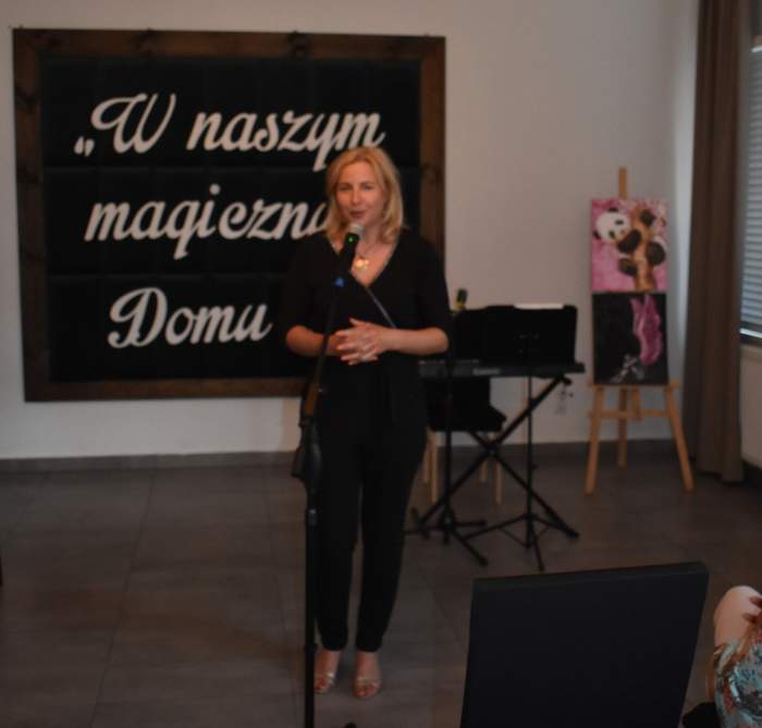 Pokaż zdjęcie: Na scenie przy mikrofonie stoi kobieta blondynka ubrana na czarno.