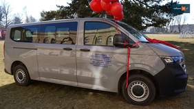 Pokaż zdjęcie: Na zdjęciu widać srebrny bus stojący bokiem do zdjęcia. Na masce i wzdłuż boku przewiązana jest czerwona wstążka, a nad dachem unoszą się czerwone balony. Na drzwiach bocznych jest nadruk z tekstem: „ Warsztaty Terapii Zajęciowej przy Stowarzyszeniu PRO-AKTIV”. Za samochodem są drzewa.