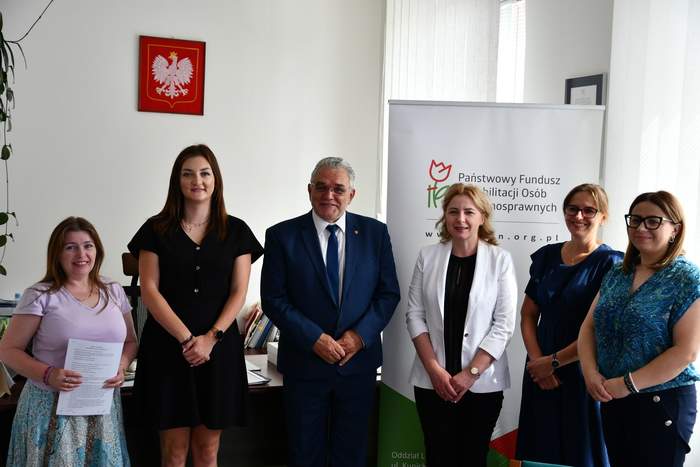Pokaż zdjęcie: Na tle roll-upu z logo PFRON pozuje do pamiątkowego zdjęcia grupa 6 osób, od lewej kobieta w spódnicy z długimi włosami, kobieta w ciemnej sukience do kolan, mężczyzna w garniturze, kobieta blondynka w białej marynarce, kobieta blondynka w ciemnej bluzce oraz kobieta w kolorowej zielonej bluzce