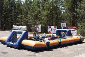 Pokaż zdjęcie: Uczestnicy Spartakiady grają na dmuchanym boisku piłkarskim, otoczonym przez banery sponsorów i organizatorów.