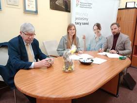 Pokaż zdjęcie: Przy podłużnym stole siedzi kobieta i dwóch mężczyzn. Zawierają umowę. Na stole leżą różne dokumenty, pieczątki, wielkanocne dekoracje. 