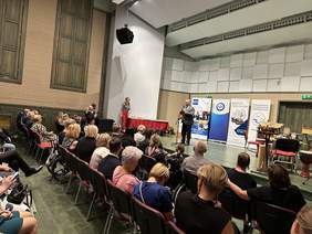 Pokaż zdjęcie: Na scenie stoi mężczyzna w marynarce, białej koszuli i krawacie. Mówi do mikrofonu, przed nim siedzą na galowo ubrani goście.