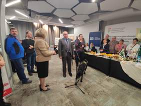 Pokaż zdjęcie: Niewidomy, elegancko ubrany mężczyzna przemawia do mikrofonu. Obok niego jest pies przewodnik.