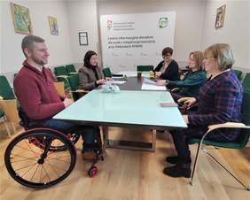 Pokaż zdjęcie: Sala konferencyjna z dużym stołem. Przy stole siedzą kobiety i mężczyźni, rozmawiają. Na stole leżą dokumenty.