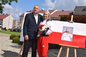 Pokaż zdjęcie: Na Rynku w Wieruszowie stoją kobieta i mężczyzna - sportowcy. Za nimi znajduje się duża flaga Polski. Pani trzyma w dłoniach biało-czerwone kwiaty. 