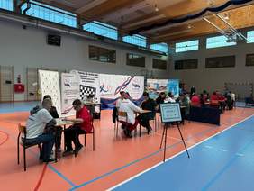 Pokaż zdjęcie: Przy trzech stołach siedzą zawodnicy, którzy grają w warcaby. Obok nich siedzą sędziowie turnieju. W tle widać banery i plakaty wydarzenia. 