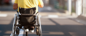 Pokaż zdjęcie: Na ulicy znajduje się ręczny wózek inwalidzki, na którym siedzi człowiek. Jest widoczny tylko jego tył. Osoba ubrana jest w żółtą koszulkę i adidasy z białą podeszwą. 