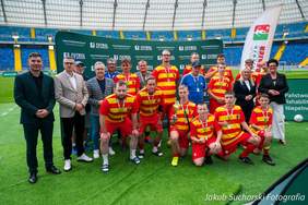 Pokaż zdjęcie: Grupa zawodników Jagiellonii Futsal „Bez Barier” w żółto-czerwonych strojach wraz z trenerami i przedstawicielami organizatorów pozuje do zdjęcia na murawie Stadionu Śląskiego.