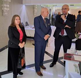 Pokaż zdjęcie: Trzy osoby, w tym jedna kobieta stoją przy stole w sali restauracyjnej i rozmawiają.