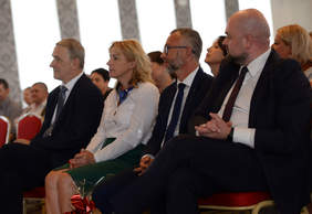 Pokaż zdjęcie: Zdjęcie sali, na krzesłach siedzą elegancko ubrani mężczyźni i kobiety