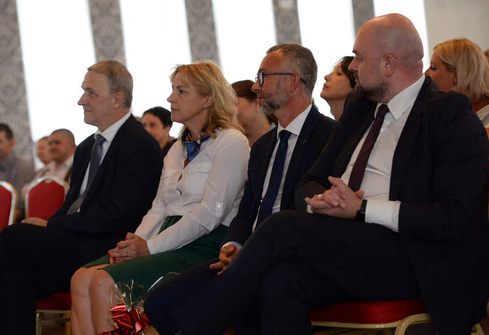 Pokaż zdjęcie: Zdjęcie sali, na krzesłach siedzą elegancko ubrani mężczyźni i kobiety