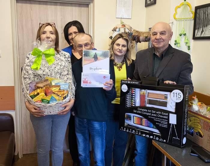 Pokaż zdjęcie: Do pamiątkowego zdjęcia pozuje pięć osób, jedna trzyma kosz cukierków, druga dyplom, kolejna zestaw plastyczny.