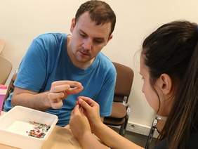 Pokaż zdjęcie: Podopieczny nawleka koralik na igłę, którą trzyma terapeuta