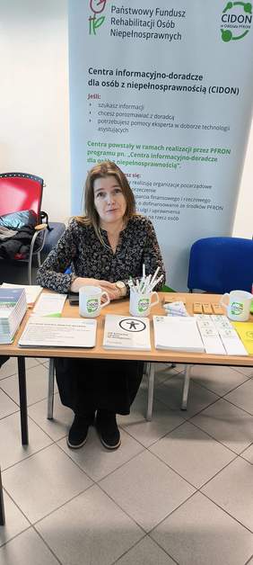 Pokaż zdjęcie: Przy stoliku na którym leżą ulotki siedzi kobieta z długimi włosami, w tle rollup z logo CIDON.