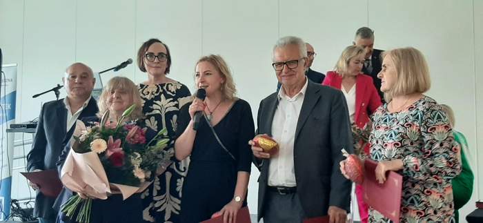 Pokaż zdjęcie: Przed scena stoi grupa sześciu osób, wraz z kwiatami, kobieta blondynka składa życzenia, trzyma w ręku mikrofon.