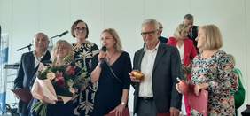 Pokaż zdjęcie: Przed scena stoi grupa sześciu osób, wraz z kwiatami, kobieta blondynka składa życzenia, trzyma w ręku mikrofon.