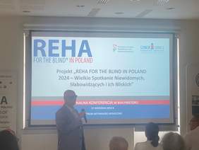 Pokaż zdjęcie: Na zdjęciu prelegent stoi przed ekranem podczas konferencji. Na ekranie widoczny jest tytuł projektu „REHA FOR THE BLIND IN POLAND 2024 – Wielkie Spotkanie Niewidomych, Słabowidzących i ich Bliskich".