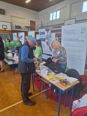 Pokaż zdjęcie: Targi pracy, na stole leżą ulotki, foldery i katalogi. Przy stole stoją dwie osoby kobieta i mężczyzna w tle roll-up z logo CIDON.