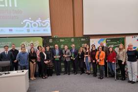 Pokaż zdjęcie: W auli na tle banerów stoi grupa ponad 20 osób do pamiątkowego zdjęcia.