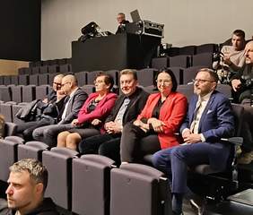 Pokaż zdjęcie: Osoby siedzące na widowni. Sześć osób w tym dwie kobiety. Oglądają występy artystyczne.