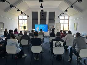 Pokaż zdjęcie: W przytulnej sali konferencyjnej na poddaszu na białych krzesłach siedzą ludzie. Patrzą na stojąca przed nimi kobietę, z mikrofonem która czyta dokument. Obok niej stoi duży wyświetlacz. Za nią duże okna.