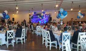 Pokaż zdjęcie: Zaproszeni goście jubileuszu 70-lecia Spółdzielni Inwalidów „Przyjaźń” siedzą w dużej sali bankietowej, na krzesłach w kolorze białym, przy stolikach przykrytych białymi obrusami. Sala udekorowana jest balonami oraz banerem okolicznościowym w tle, informującym o niniejszej uroczystości. W oddali stoi kobieta wypowiadająca się do mikrofonu.