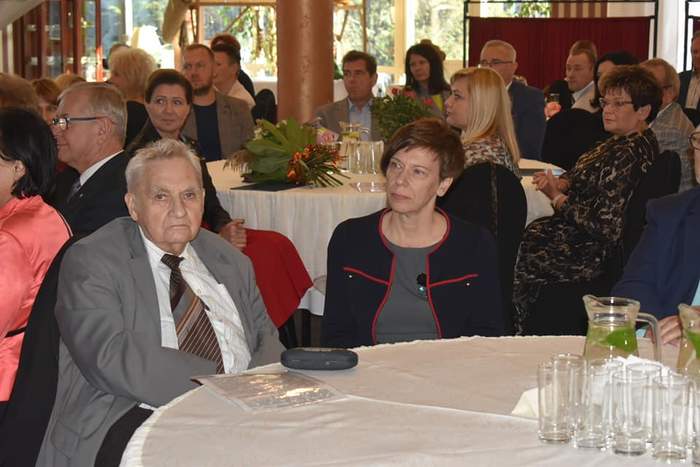 Pokaż zdjęcie: Duża grupa elegancko ubranych kobiet i mężczyzn siedzi w restauracji przy okrągłych stolikach przykrytych białymi obrusami. Na stolikach ustawione zostały szklanki oraz dzbanki z wodą. Na jednym ze stolików widoczne są dwa bukiety kwiatów.