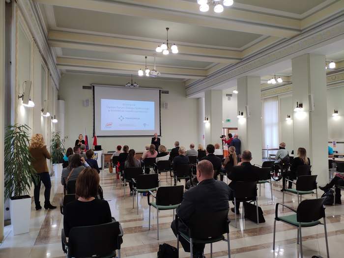 Pokaż zdjęcie: duża sala z kolumnami ludzie siedzą w rzędach pod ścianą stoi kobieta ubrana na czarni i trzyma mikrofon obok stoi kobieta i tłumaczy na język migowy