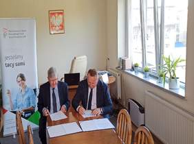 Pokaż zdjęcie: na zdjęciu pomieszczenie biurowe na ścianie widać Godło Polski z lewej strony rozmieszczono baner PFRON, na środku duży stół przy którym siedzą dwie osoby w garniturach  i podpisują dokument o wzajemnej współpracy pomiędzy PFRON a Łódzką 