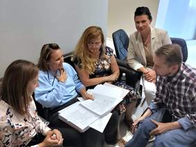 Pokaż zdjęcie: Pięcioosobowa grupa podczas spotkania roboczego. Uczestnicy siedzą w kręgu, analizują dokumenty i prowadzą rozmowę w przyjaznej atmosferze