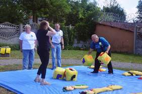 Pokaż zdjęcie: Na trawie rozłożona jest duża niebieska mata, na niej stoją 4 osoby, jedna z nich przenosi duże żółte kostki