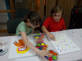 Pokaż zdjęcie: Dziewczynki podczas zajęć plastycznych - dwie dziewczynki w trakcie malowania farbami. Siedzą przy stoliku i malują palcami